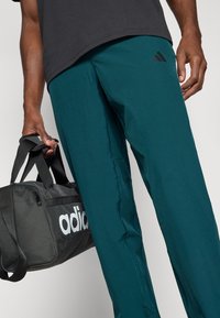 Tirkizne atletske hlače s crnim Adidas logom, uparene s sivom torbom za teretanu koja ima bijeli Adidas logo, koju osoba drži u ruci.