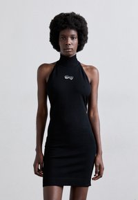 Robe sans manches en tricot noir avec un col montant. Présente une silhouette ajustée et un ourlet côtelé. Design d'emblème en argent notable sur la poitrine.