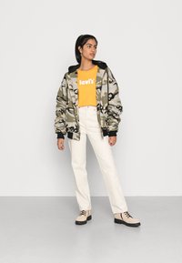 Jardineira com padrão de camuflagem, t-shirt cropped laranja com o logótipo "levi's", jeans brancos retos, botas bejes com solas pretas, em pé contra um fundo branco.