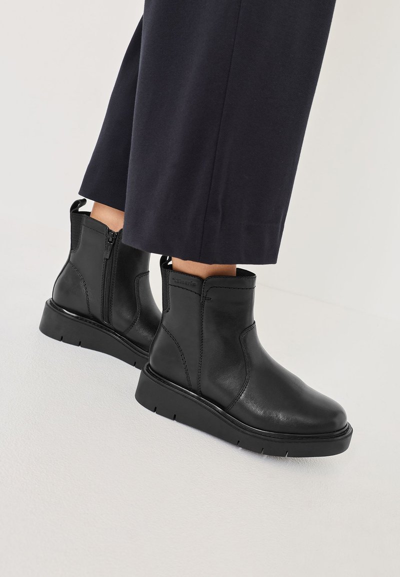 Schwarze Lederankelstiefel mit flacher Sohle, seitlichem Reißverschluss und Zuglasche; glatte Textur mit Nähdetails und minimalistischen Design.