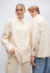 Κρεμ πουκάμισα με υφή και φθαρμένο σχέδιο, με γιακά με κουμπιά και ορατές ραφές, φορεμένα από δύο μοντέλα με ανοιχτόχρωμο φόντο.