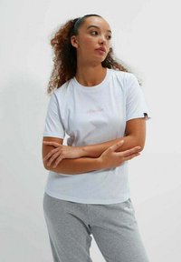 Camiseta blanca de manga corta con cuello redondo y logo rosa en el centro. Combinada con pantalones de chándal grises para un look casual.