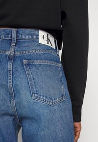 High-waisted blue denim jeans featuring a classic cut, back pockets, and a white Calvin Klein logo patch on the waistband.