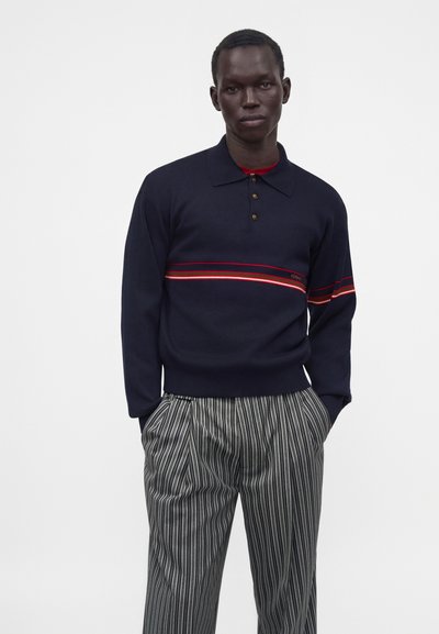 Homme portant un polo bleu marine à manches longues avec des rayures horizontales rouges et un pantalon gris à rayures verticales, debout les mains dans les poches sur fond blanc.