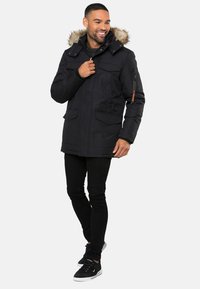 Černá parka s kapucí lemovanou kožešinou, několika předními kapsami a boční kapsou na zip. Nosené s černými džíny a tmavými teniskami.
