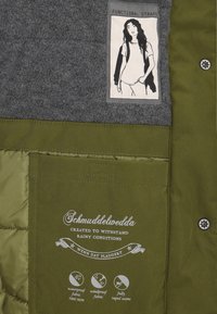 Olivgrön jacka med insida av grått tyg, med en etikett som har en illustration av en kvinna, vattentäta detaljer och funktionella remmar.