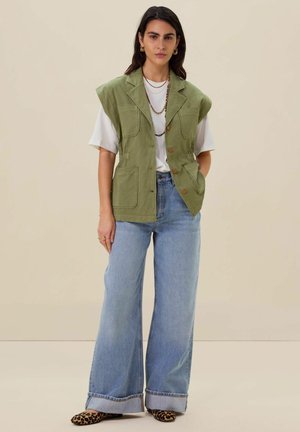 Green sleeveless vest with large pockets, button closure, worn over a white shirt and paired with wide-leg light blue jeans and animal print shoes.