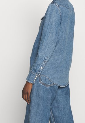 Person iført en blå denimskjorte med perlesnapmanchetter og matchende blå vidde denimbukser mod en ensfarvet baggrund.