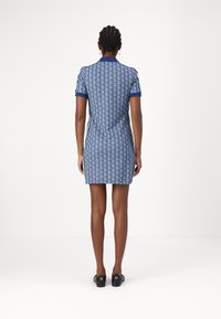Robe bleue à motifs avec une silhouette ajustée, des manches courtes et un col. Présente un design de logo répétitif et des manches contrastantes. Chaussures noires portées.