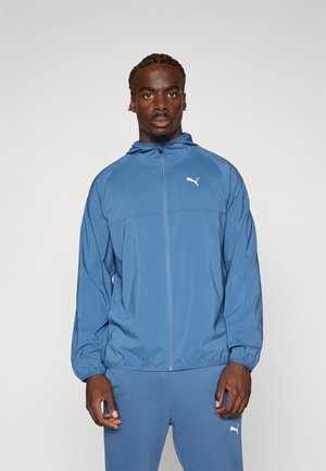 Puma windbreaker in blue material with a front zipper, hood, and elastic cuffs. Features a white logo on the chest. Paired with matching pants.