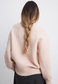 Ljusrosa stickad tröja med avslappnad passform, ribbat avslut och sänkt axel, bärs över en mörkgrå klänning. Lång flätad hår synligt.