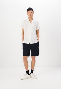 Kortärmad vit stickad skjorta med mönster, kombinerad med mörka shorts och vita sneakers. Modellen står framför en enfärgad vit bakgrund.