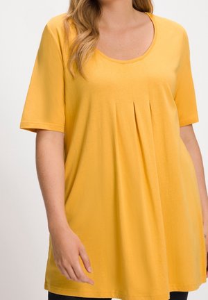 Mujer vestida con un vestido amarillo de manga corta y holgado, con cuello redondo y pliegues delanteros, recortado en los hombros y a mitad del muslo.