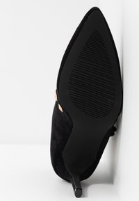 Chaussure à talon haut en daim noir avec un bout pointu et un talon fin. La semelle est texturée avec des rainures horizontales. Accents en métal doré visibles.