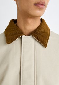 Chaqueta beige con un amplio cuello de pana marrón. Presenta un diseño limpio y costuras visibles. La tela parece suave y resistente.