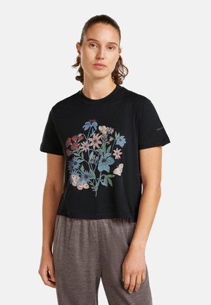 Kvinde med sat hår tilbage iført en sort t-shirt med farverigt blomster- og sommerfugledesign, kombineret med løstsiddende grå bukser.