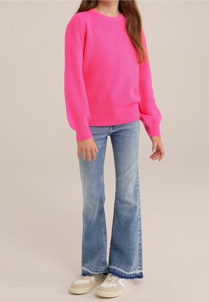 Person iført en lyserød tekstureret sweater, lyseblå flare-jeans og beige sneakers stående mod en ensfarvet beige baggrund.