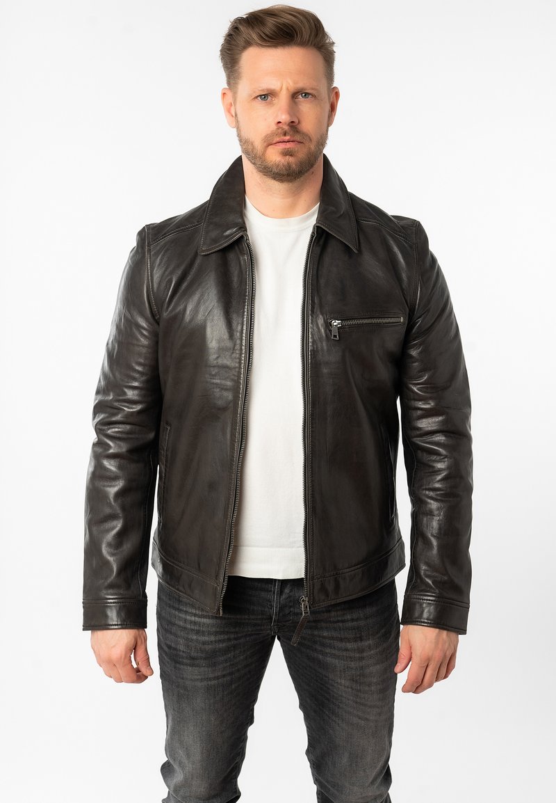 Zwarte leren jas met een gladde textuur, voorzien van een puntige kraag, een ritssluiting aan de voorkant en een borstzak met rits. Draagt over een wit shirt.