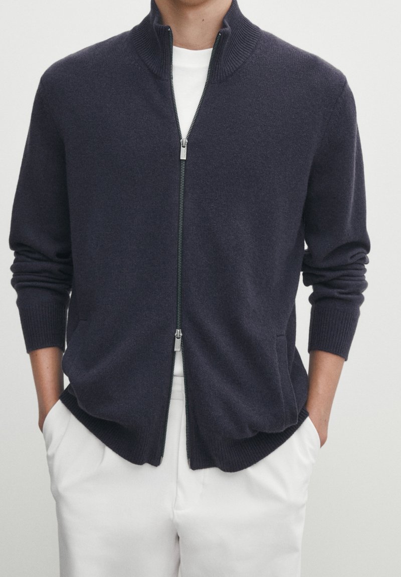 Homme portant un pull zippé bleu marine foncé sur une chemise blanche, les mains dans les poches d’un pantalon blanc, sur un fond uni.