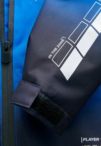 Blaue Jacke mit schwarzen Ärmeln, ausgestattet mit einem Klettverschluss. Weißer Text "IN THE ZONE" und geometrische Muster am Ärmel. Wasserabweisendes Material.