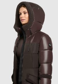 Padded brown jacket with a plush, fur-lined hood, front zipper, and side pockets. Features a shiny finish and contrasting textured accents.