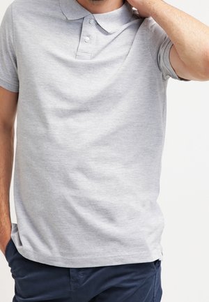 Uomo che indossa una polo a maniche corte grigio chiaro e pantaloni blu scuro, una mano in tasca, l'altra mano che tocca il collo.