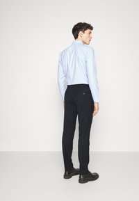 Camicia azzurro chiaro a maniche lunghe con colletto button-down, abbinata a pantaloni scuri. La vista posteriore mostra un design aderente e dettagli della tasca. Scarpe nere.