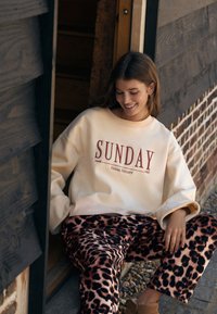 Lichtbeige sweatshirt met rode "SUNDAY" print, gecombineerd met roze en zwarte broek met luipaardprint. Losse pasvorm, gestructureerde stof.
