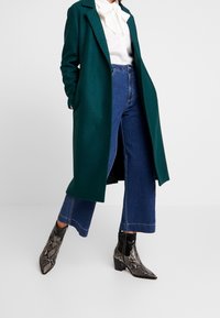Manteau long vert en tissu texturé, associé à un chemisier blanc et à un pantalon en denim bleu taille haute évasé. Bottes a imprimé serpent noir et argent.