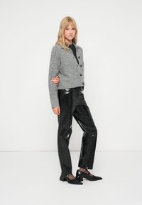 Noor blondide juustega noor naine kannab halli nööpidega kardigani, musti läikivaid nahkpükse ja musti madala kontsaga kingi, seistes valge tausta ees.