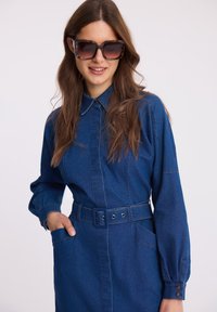 Dżinsowa sukienka z długimi rękawami, detalem paska i mankietami zapinanymi na guziki. Głęboki niebieski kolor z kontrastowym stębnowaniem i bocznymi kieszeniami. Okulary przeciwsłoneczne w kolorze żółwia.