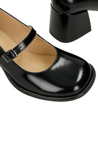 Czarny lakierowany but typu Mary Jane na masywnym słupku z regulowanym paskiem na klamrę, beżowa wyściółka wewnętrzna i zaokrąglony nosek.