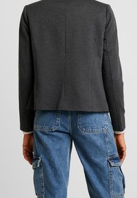 Blazer gris texturé à coupe raccourcie, avec une couture dos nette, associé à un pantalon cargo bleu et des poignets côtelés visibles.