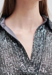 Schwarzes Paillettenshirt mit V-Ausschnitt und grauem Kragen, mit einer strukturierten Oberfläche durch die glänzenden Pailletten, die das Licht aus verschiedenen Winkeln reflektieren.