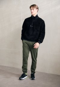 Sort fleece halv-zip trøje, grøn sweatpants med siden tekst, og sorte sneakers med neonlys. Blød tekstur og afslappet pasform.