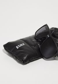 Schwarze Sonnenbrille mit quadratischen Rahmen, dunklen Gläsern und Logodetails, die auf einem schwarzen gepolsterten Etui mit leicht glänzender Textur liegt.
