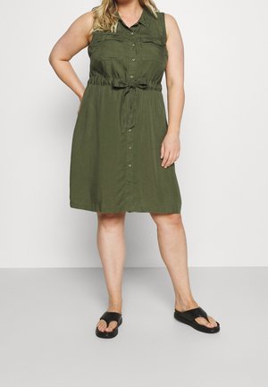 Kobieta ubrana w bezrękawową oliwkowo-zieloną sukienkę zapinaną na guziki z paskiem w talii oraz czarne sandały z odsłoniętymi palcami, stojąca na jednolitym tle.