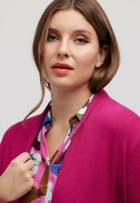 Fuchsia Cardigan, weiches Strickmaterial, offenes Design, über einer bunten, blumenmusternden Bluse mit verschiedenen Farbtönen und Formen getragen.