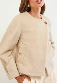 Vrouw in een beige wollen jas met oversized mouwen, gouden decoratieve knopen en zijzakken, gecombineerd met lichtgekleurde broek.