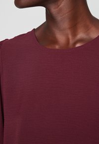 Haut bordeaux avec un col rond, une texture douce et un léger froncé à l'épaule. Design décontracté avec une finition lisse.