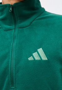 Gros plan d'une veste polaire verte avec un zip quart et un logo brodé blanc à trois barres sur le côté gauche de la poitrine.