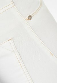 El tejido de denim blanco presenta costuras visibles y un acento de botón metálico redondo cerca del bolsillo. Textura suave y construcción robusta.