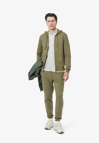 Felpa verde oliva sovrapposta a una maglietta a righe, abbinata a pantaloni jogger coordinati e sneakers bianche. La felpa presenta una tasca frontale e cordini.