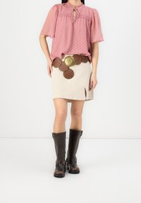 Blouse rose à pois avec des manches bouffantes, mini jupe beige avec fente sur le côté, ceinture marron avec de grands accents circulaires et bottes marron jusqu'aux genoux.