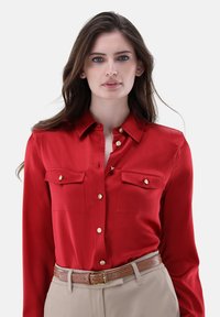 Rotes Seidenhemd mit zwei Brusttaschen, goldenen Knöpfen und einem Button-Down-Kragen, kombiniert mit beigefarbenen Hosen und einem strukturierten braunen Gürtel.