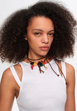 Mujer con cabello rizado que lleva una camiseta blanca sin mangas y un collar de múltiples colores con forma de rama atado con cordones marrones.