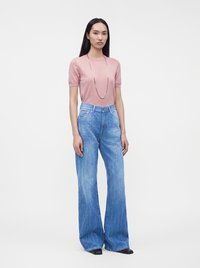 Femme aux longs cheveux noirs portant un haut rose à manches courtes, un long collier, un jean bleu taille haute à jambes larges et des chaussures noires, debout sur un fond blanc.