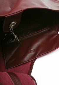 Maroon leather handbag interior featuring a zippered pocket and soft suede texture, with a visible logo embossed on the lining.