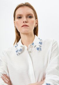 Camicia bianca con tessuto a trama e colletto caratterizzato da motivi floreali ricamati in blu e bianco. Chiusura con bottoni sul davanti.