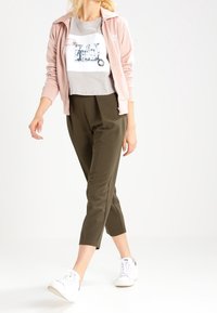 Rosa zip-up jacka över en grå T-shirt med grafiktryck, i kombination med olivgröna tapered byxor och vita sneakers. Slät tyg, avslappnad design.